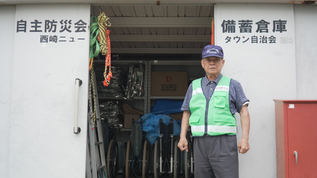 備蓄倉庫の前に立つ古我知進さん。倉庫には『自主防災会 備蓄倉庫 西崎ニュータウン自治会』の表記