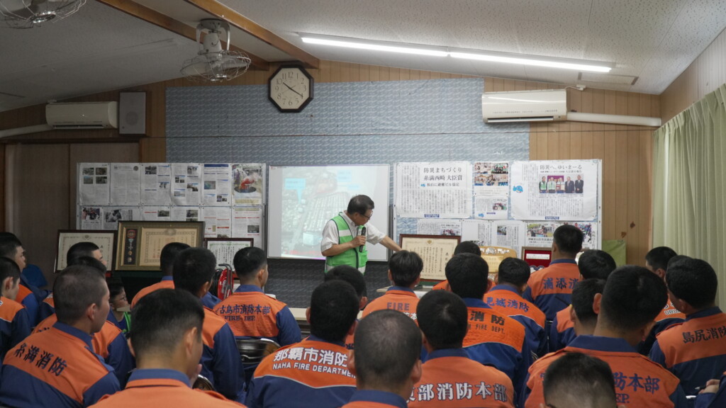 沖縄県消防学校の初任科学生に講演する古我知進さん。スクリーンには西崎ニュータウンの航空写真が映し出されている
