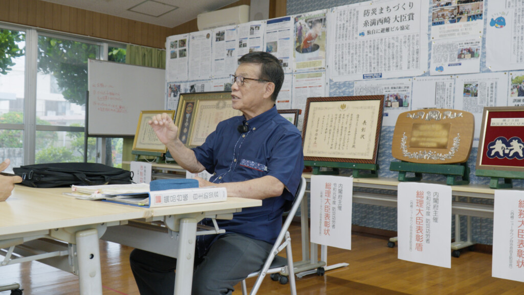 取材に応じる古我知進さん。背後には数々の表彰状と新聞記事が並ぶ