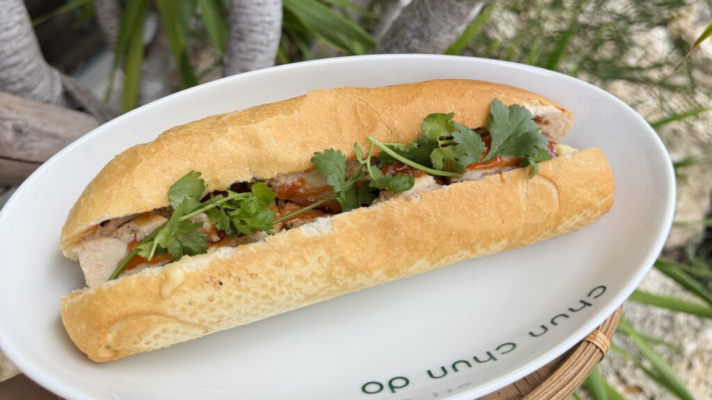 グリルチキンバインミー。パクチーとチリソースがかかったベトナムサンドイッチが、観葉植物を背景に白い皿に盛られている