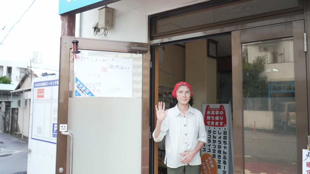 お店の入り口でお客さんを見送る、割烹着姿の稲嶺さん。ガラス扉越しに笑顔で手を振っている