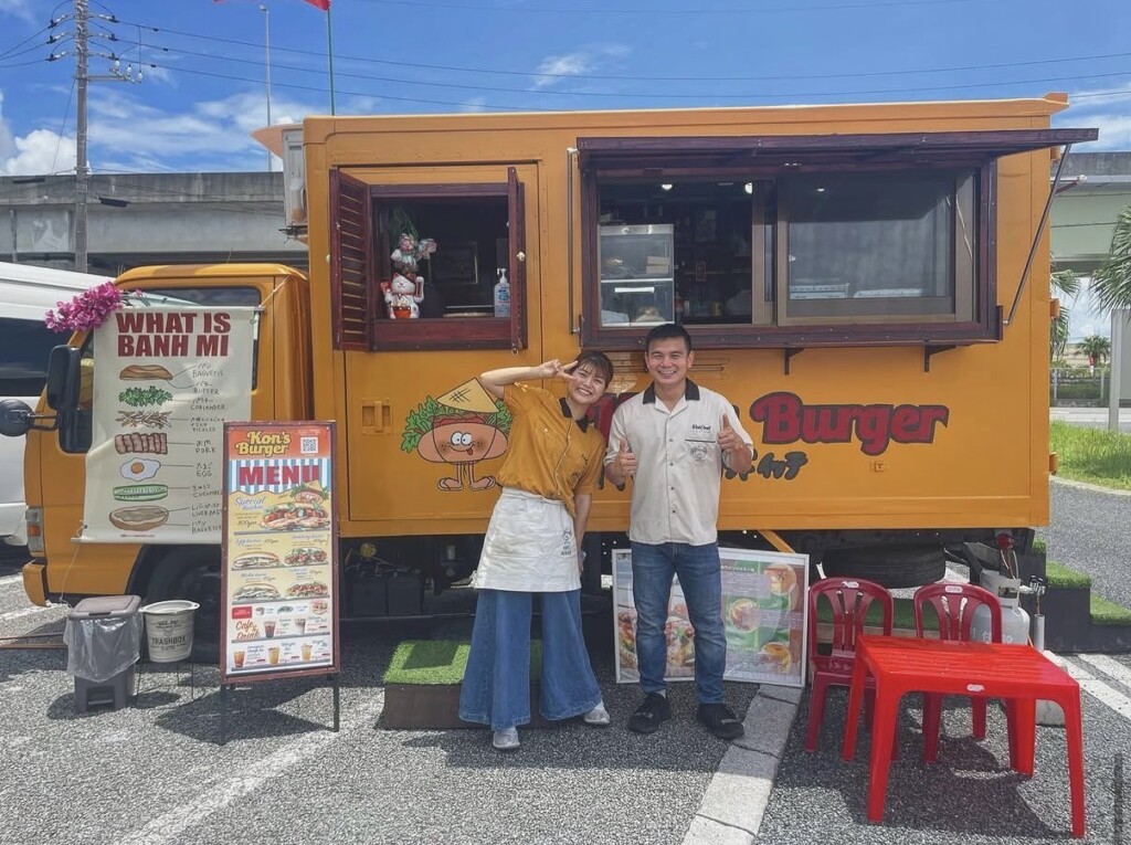オレンジ色のキッチンカーの前に立つコンさんと奥さん。車体には『WHAT IS BANH MI』のポスターとメニュー、Kon's Burgerのロゴが見える