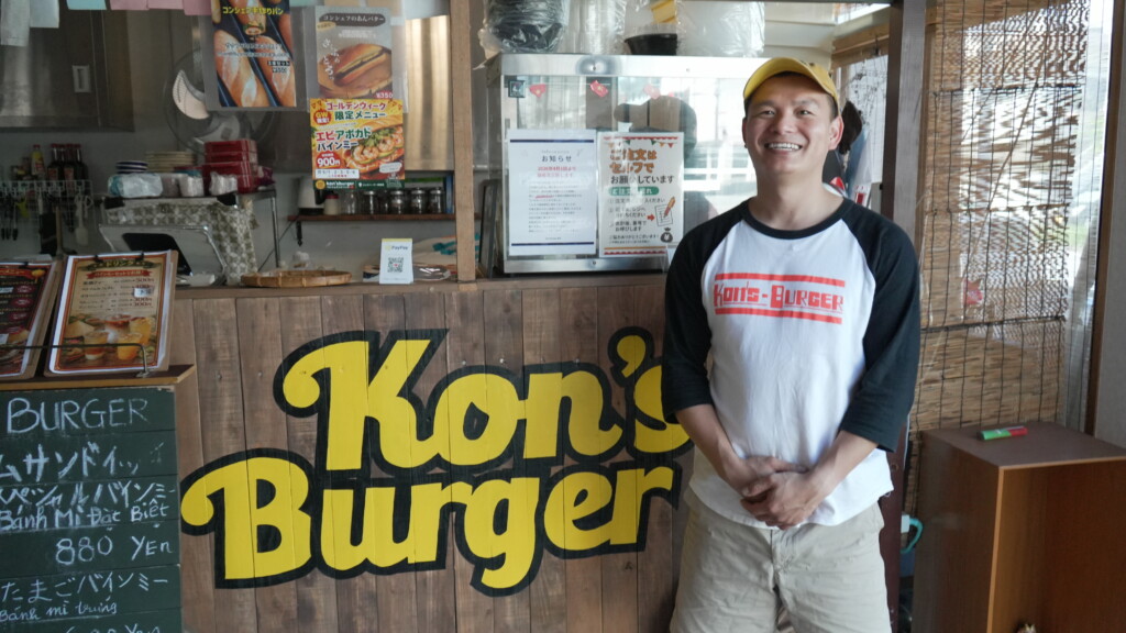 コンズバーガーのオーナー、コンさんが店内のKon's Burger看板の前で笑顔で立っている。糸満市西崎のベトナムサンドイッチ専門店