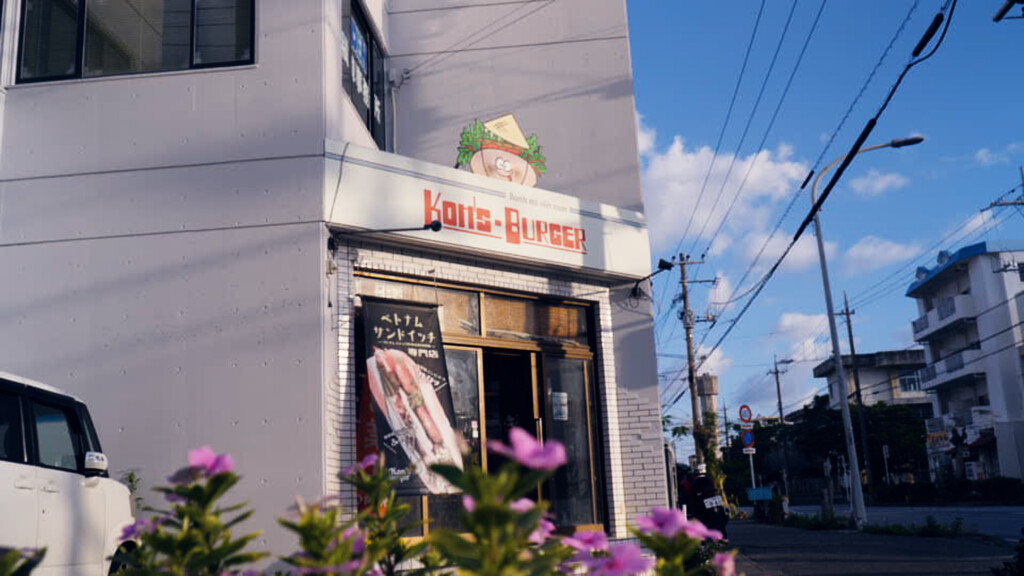 コンズバーガー西崎店の外観。白いビルの1階に赤い文字でKON'S BURGERの看板。手前に紫の花が咲いている