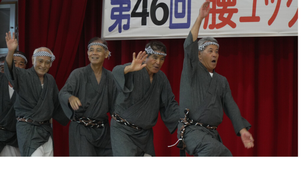 赤幕の舞台で、鉢巻きに作務衣姿で笑いながら群舞するゲキラン十日の会のメンバー（腰ユックイ行事）