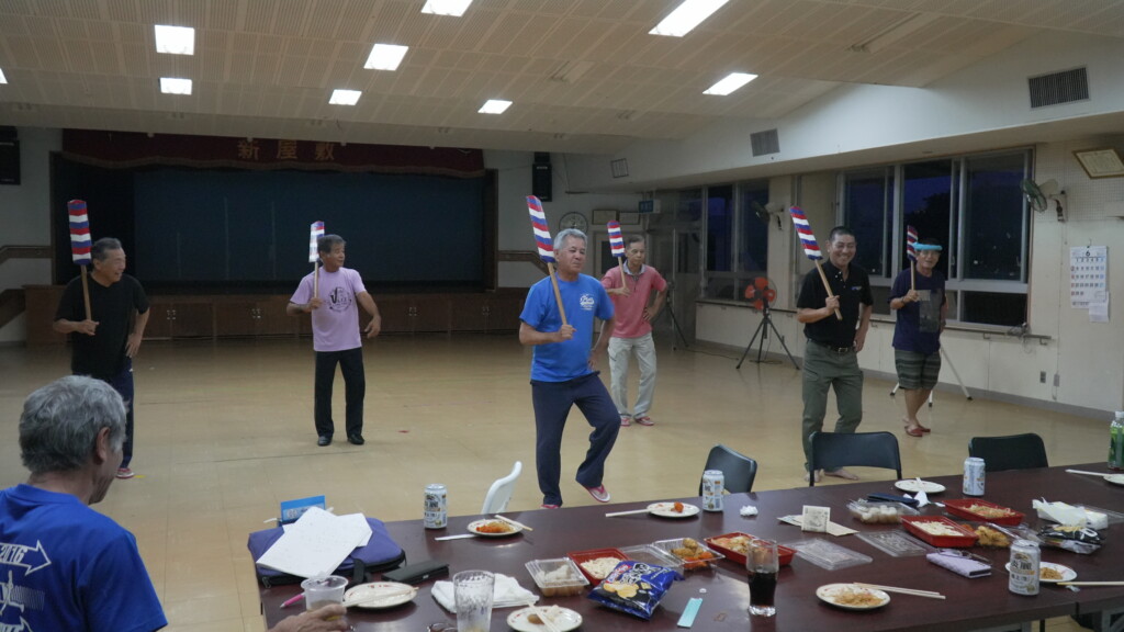 新屋敷区自治会館での水曜夜の練習。私服のメンバーがエークを持ち、手前のテーブルに酒とつまみが並ぶ